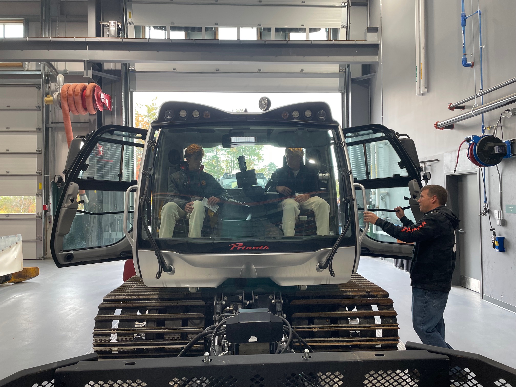 Two young people sit in the cab of a Prinoth snow grooming machine with its doors open inside a maintenance garage, while a man standing alongside shows them the controls.